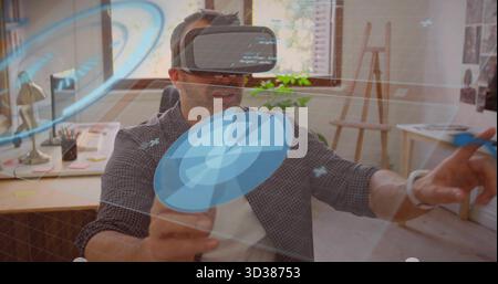 Interacting mature man wearing virtual reality headset sitting in studio with desk lamp and easel Stock Photo