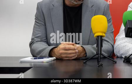 An unrecognizable man with his fingers intertwined is sitting in front of a table with several microphones. He appears relaxed and professional Stock Photo