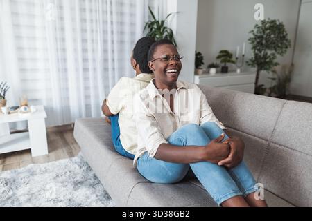 Two cheerful african american women relaxing on sofa at home, laughing and enjoying time together Stock Photo