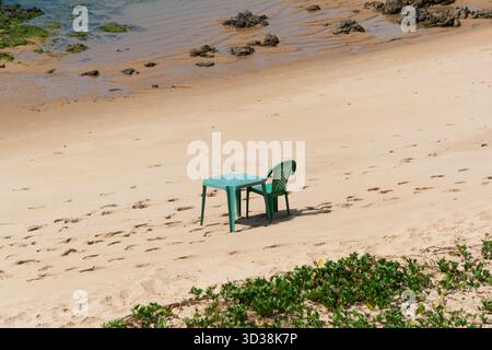 A tranquil and sunny beach scene, with a table and two colorful plastic chairs arranged on the beige sand. Stock Photo