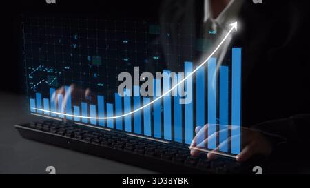 A business professional interacts with a keyboard while overseeing a digital graph, illustrating growth and success in a dark-themed workspace focused Stock Photo