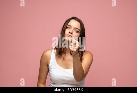Confident emotion of young woman pointing finger forward in white tank top on pink background Stock Photo