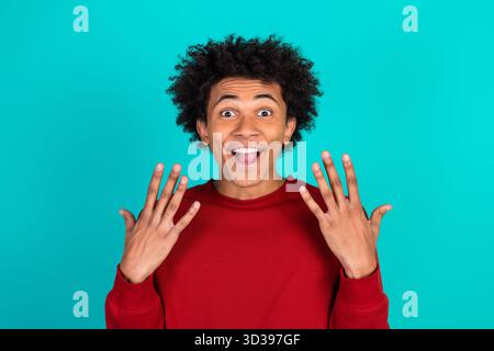 Cheerful young man in red sweater shows excited expression with open hands against a vibrant teal background Stock Photo