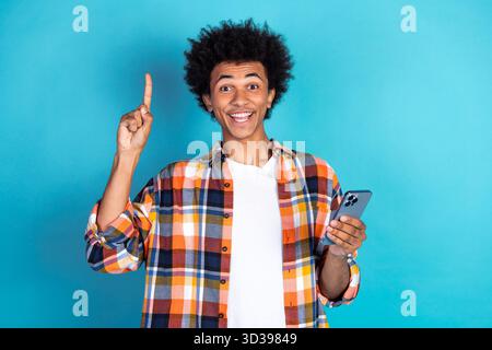 Young male student smiles and points up with a smartphone in a casual colorful shirt against a bright blue background Stock Photo