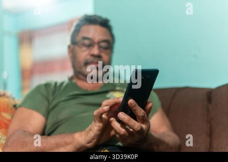 A side profile close-up of a middle-aged Asian man with a beard and glasses, sitting on a sofa and intently playing a mobile game on his smartphone. Stock Photo