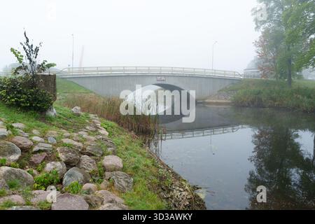 An atmospheric horizontal shot of a modern concrete bridge reflected in the still water of a river on a dense, misty morning. The image conveys calm, Stock Photo