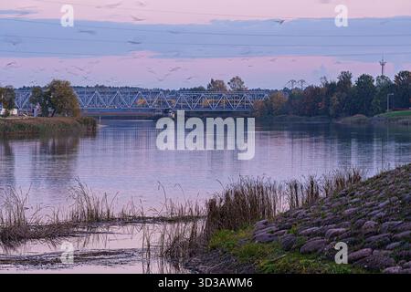 A serene landscape featuring a steel railway truss bridge spanning a calm river under a soft, colorful sunset sky, with autumn trees and waterfront ve Stock Photo
