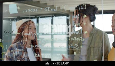 Women in varied attire collaborating on code behind glass board in office, with pastel sticky note Stock Photo
