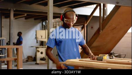Sanding carpenter wearing goggles and earmuffs using power sander at workshop, wooden planks Stock Photo