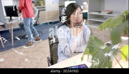 Talking Korean woman using smartphone while viewing laptop in workspace, with currency symbols Stock Photo