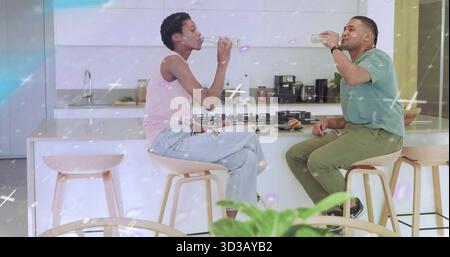 Two friends drinking glass water bottles in home kitchen with island, with light wood bar stools Stock Photo