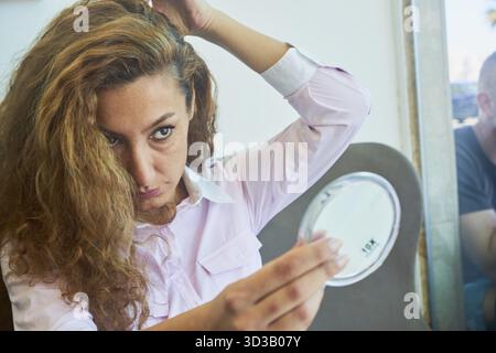 Mature woman examining her face in mirror, long brown curly hairs, self-perception, handglass, hand mirror, checking appearance of herself, middle age Stock Photo
