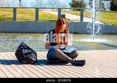 In a vibrant park setting, a young woman sits by a fountain, focused on her studies. The sun shines brightly, creating a serene atmosphere as she writ Stock Photo