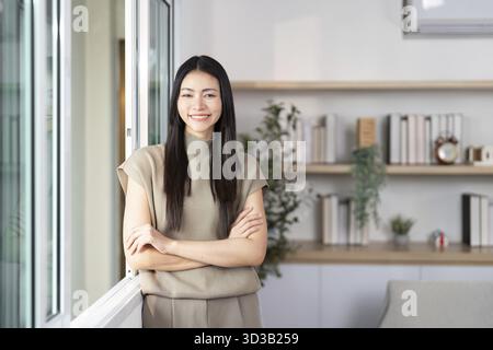 A confident woman stands by the window, smiling warmly in her stylish home environment Stock Photo