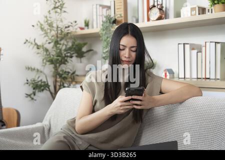 A focused young woman engages with her smartphone while sitting comfortably on a couch in a cozy home environment Stock Photo