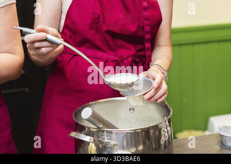 Food, Close-up of a woman in red apron using a ladle to pour creamy batter into a foil cup from a large pot Stock Photo