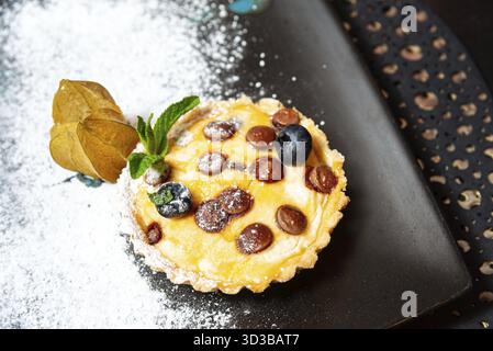 Food, Close-up of a baked mini tart topped with chocolate drops, blueberries, and mint, served on a black plate with powdered sugar Stock Photo