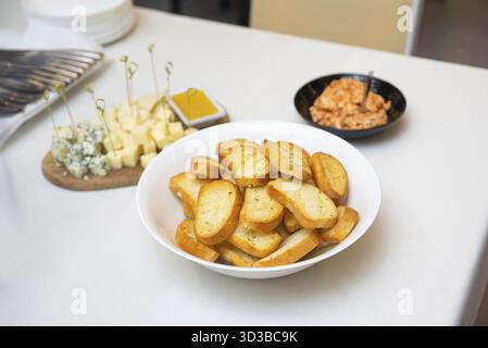 Food, Crispy toasted baguette slices served in a bowl, accompanied by a cheese platter and dipping sauces on a white table Stock Photo
