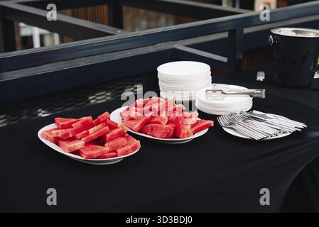 Food, Freshly cut watermelon slices arranged on plates beside utensils and stacked dishes on a black tablecloth at an outdoor event Stock Photo