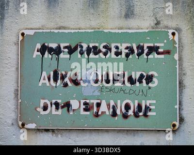 Sare, France - Oct 09 2025: Weathered petanque sign defaced with Basque language activist graffiti, captured on a wall in Sare, Pyrenees-Atlantiques, France. Stock Photo