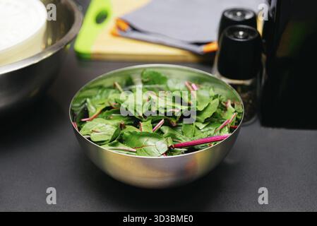 Food, A stainless steel bowl filled with fresh Swiss chard leaves with red stems, placed on a dark kitchen countertop near cooking tools Stock Photo
