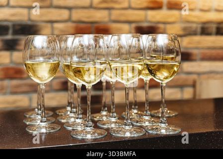 Food, A collection of elegant wine glasses filled with white wine, arranged on a bar counter against a rustic brick wall background Stock Photo