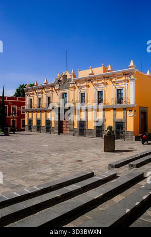 Vibrant orange and yellow colonial building in a plaza, featuring black volcanic stone stairs and a deep blue sky in Mexico. Stock Photo