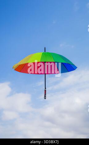 Vertical shot of a multicolored umbrella with rainbow colors floating in the air against a clear blue sky background with white clouds. Stock Photo