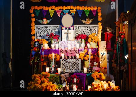 Front view of a richly decorated Day of the Dead altar with roses, marigold, white cutout papel picado, and lit candles against a dark background. Stock Photo
