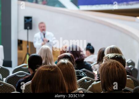 Crowd of attendees listens to speaker delivering talk at modern venue. Audience sits in rows, stage behind, equipment and banners visible, event atmosphere focused and professional. Stock Photo