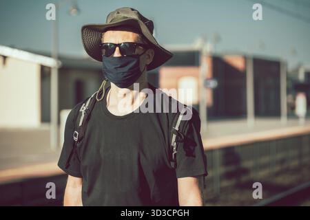 Middle aged male man wearing protective face mask, standing alone on an empty platform awaiting arrival of a scheduled train, traveling in the time of Stock Photo