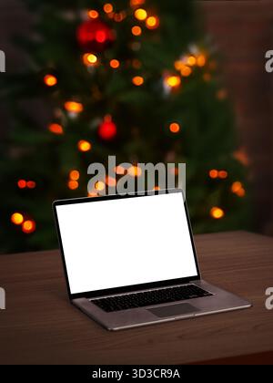 Open laptop with blank screen on wooden table, Christmas tree lights glowing in soft focus background, cozy festive atmosphere Stock Photo