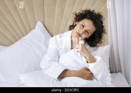 Sad depressed curly woman hugging pillow sitting in white bed alone Stock Photo