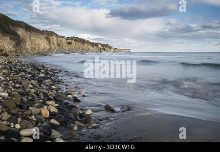 Ocean waves crashing on a rocky beach with cliffs in the background on a cloudy day. Cyprus Stock Photo