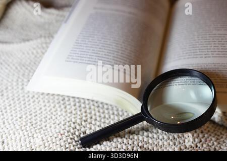Open book with a magnifying glass resting on a knitted blanket, emphasizing detailed reading, analytical study, and the pursuit of knowledge in a cozy Stock Photo