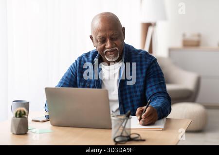 Senior Black Man Takes Notes While Working on Laptop at Home During Daytime Stock Photo