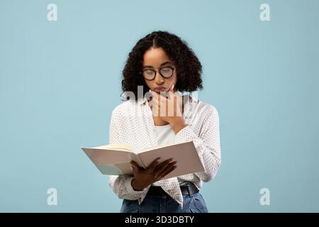 Pensive Black Female Student Reading Textbook While Studying for Exam Against Blue Background Stock Photo