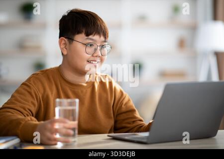 Healthy Asian Teenager Enjoys Water While Studying at Home on a Laptop Stock Photo
