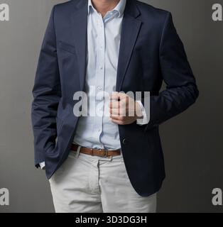 Man standing in a smart suit against a grey wall holding one side of his jacket Stock Photo