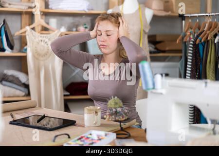 Tired female fashion designer sitting at table in sewing studio Stock Photo
