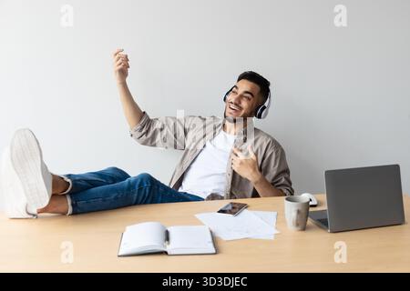 Joyful Arab Man Relaxing at Home Office While Playing Virtual Guitar and Listening to Music Stock Photo