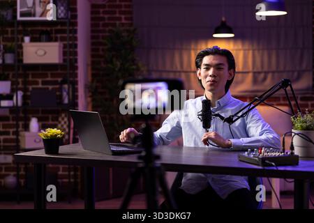 Modern asian podcaster works at desk with microphone and laptop, creating digital audio programs. Male vlogger delivers content aimed at online branding, social media strategy and creative businesses. Stock Photo