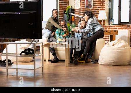 Happy friends gather in living room, raising beer bottles, laughing and enjoying snacks during casual hangout. Smiling diverse group sit on couch, watching tv and having drinks in apartment. Stock Photo