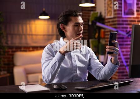Focused asian man working late at home, using smartphone for video call with colleagues in different time zone. Businessman shares strategy ideas and gestures while discussing project details remotely Stock Photo
