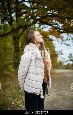 A young woman enjoys a peaceful moment outdoors, embracing the autumn air with a serene smile while surrounded by vibrant foliage. Stock Photo