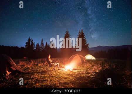Hiker woman relaxes by campfire, illuminated by its warm glow, with tent nearby under stunning, star-filled sky. Milky Way stretches overhead, adding magical touch to serene mountain setting. Stock Photo