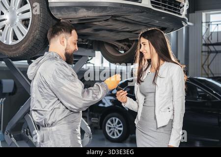 Shot of Auto Mechanic giving the car keys to his female customer in workshop at auto repair center. Quality of Service, Customer Satisfaction Stock Photo