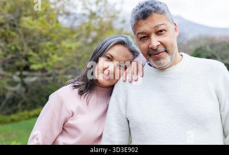 Senior couple in sweaters standing close together looking at camera in grassy yard under cloudy sky Stock Photo