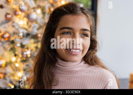 Child girl wearing pink sweater gazing at Christmas tree with fairy lights at home near window Stock Photo