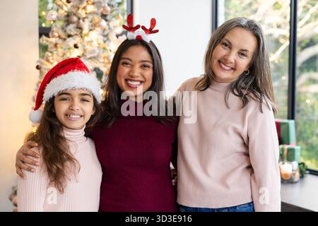 Diverse female family standing at home by decorated Christmas tree wearing sweaters and headgear Stock Photo